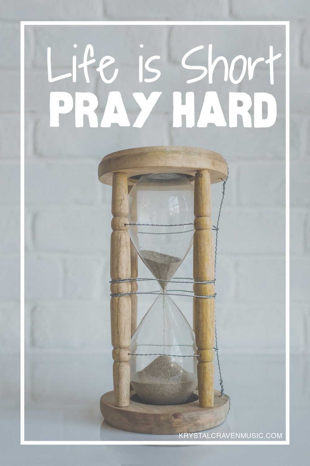 The words, "Life is short, pray hard" above an hourglass with a wooden frame on a white counter with a white brick backsplash.