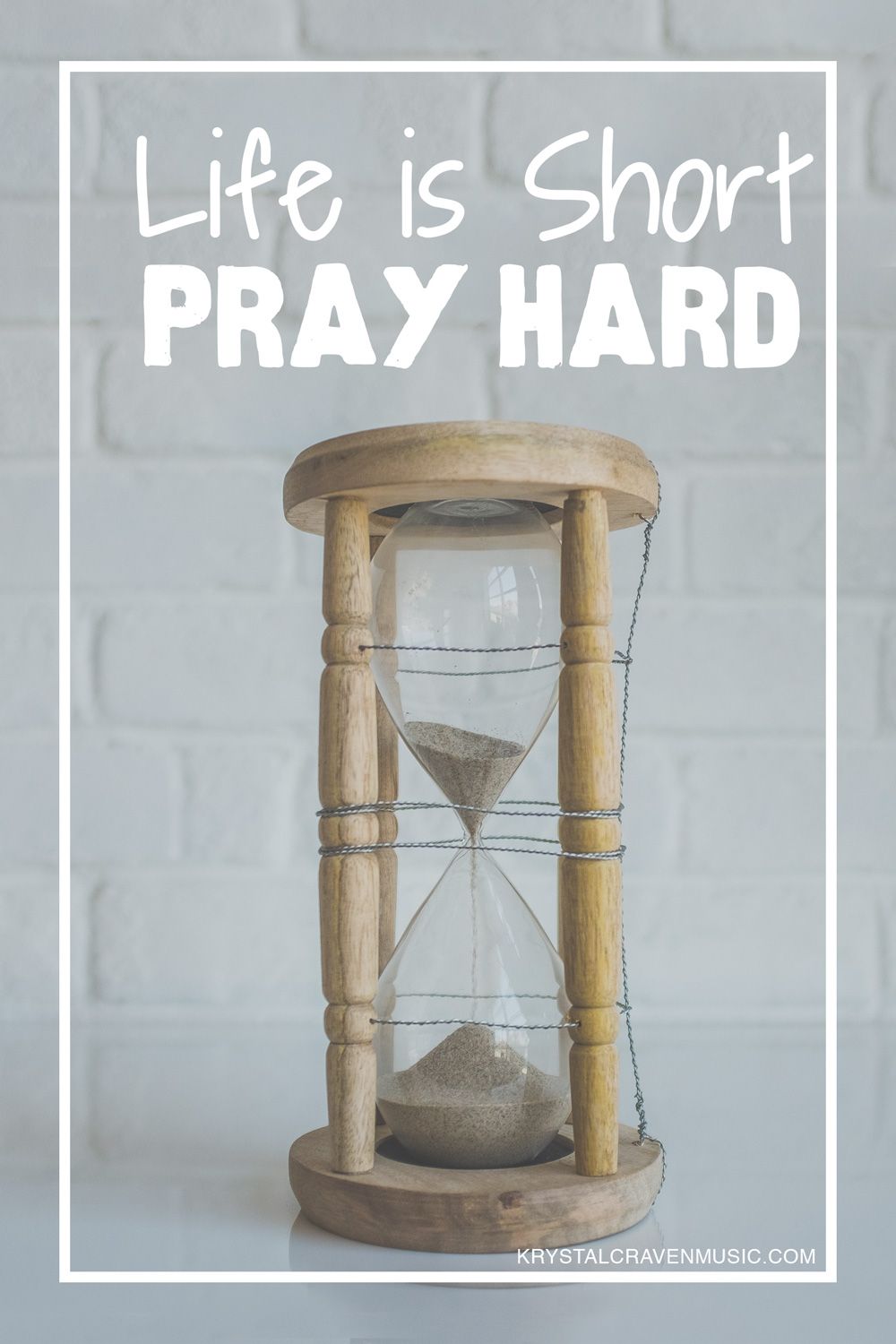 The words, "Life is short, pray hard" above an hourglass with a wooden frame on a white counter with a white brick backsplash.