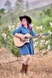 Krystal outside with flowers and mountains in the background. She's playing a guitar and is wearing a black cowboy hat, brown cowboy boots, and a denim dress