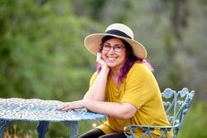 Krystal smiling outside at a bistro table with her head propped on her hand wearing a yellow shirt and straw colored hat