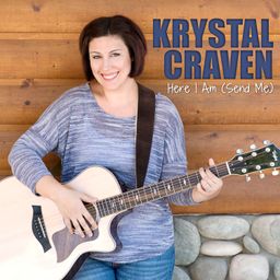 Krystal standing in front of a log wall holding her guitar and looking at the camera. Above her left shoulder is her name in blue text with the title 'Here I Am (Send Me)' in white text below it.