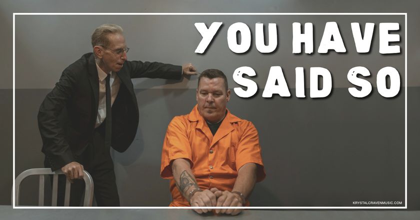 A man in an interrogation room in an orange jumper seated with his hands cuffed together on a table while a man standing next to him is leaning towards him and speaking with his fist against the wall with the words "You Have Said So" overlaying the image.
